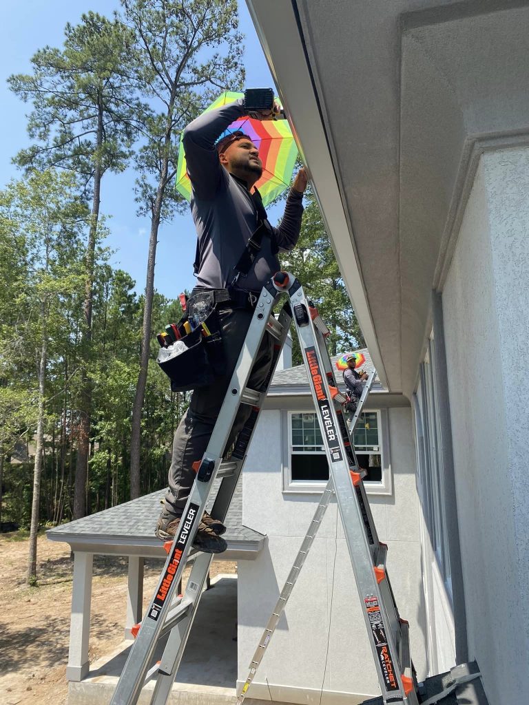 Worker on a ladder installing a gutter system along a roofline, wearing tool gear and working outdoors near trees