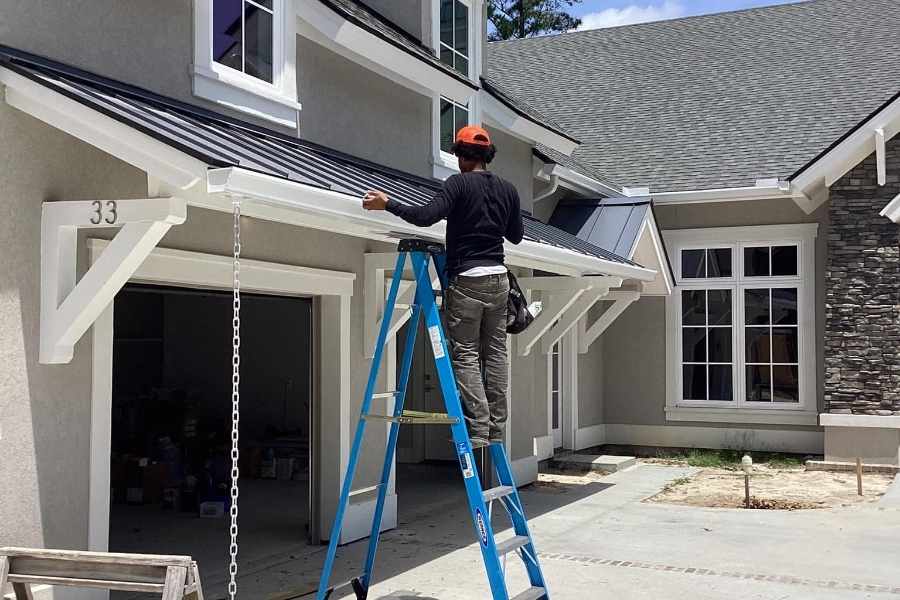 Technician on a ladder installing a gutter system along the roofline of a home with white trim and metal roofing accents