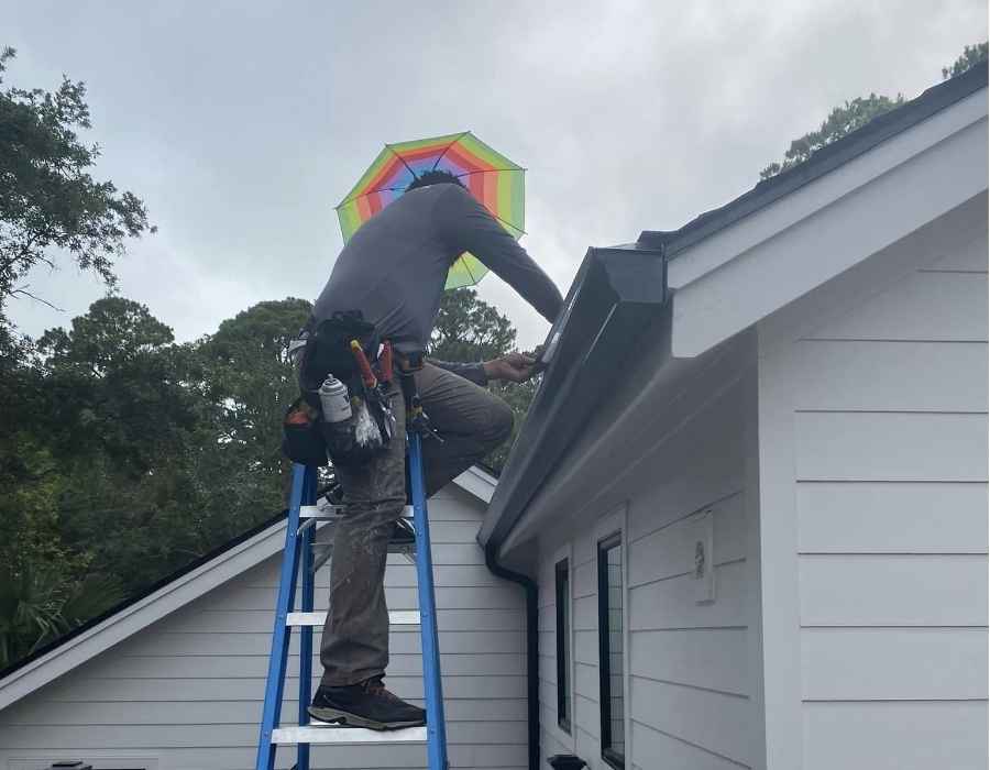 Technician standing on a ladder working on a gutter system, wearing tools and using an umbrella for sun protection