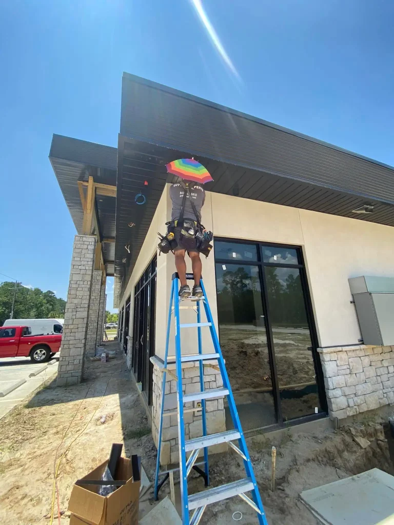 A gutter installer is standing high up on a tall blue A-frame ladder, working on a long stretch of dark horizontal metal gutters that has just been installed. He is attached to a safety harness that runs down to his large, fully-stocked tool belt. The entire scene is drenched in extremely bright, direct sunlight, creating an harsh glare against the blue sky and sharp shadows. The sun is beaming so intensely that even with the colorful rainbow shade overhead, the area he is working on is brilliantly illuminated. He is meticulously securing the dark gutter to the roofline, which sits above the new building's stucco and stone-textured walls. Below him, the ground is dirt and construction materials are scattered nearby, including a cardboard box. In the background, there are trees and a red pickup truck.