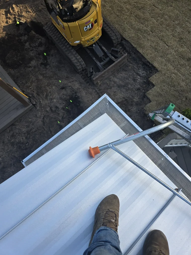 A first-person, high-angle view from a metal roof showing the professional installation of stainless steel micro-mesh gutter guards. A worker's boots are visible on the white standing-seam roof near a ladder stabilizer. Below on the ground, a yellow Caterpillar mini-excavator is parked near a freshly excavated trench, demonstrating a coordinated new construction or drainage site.