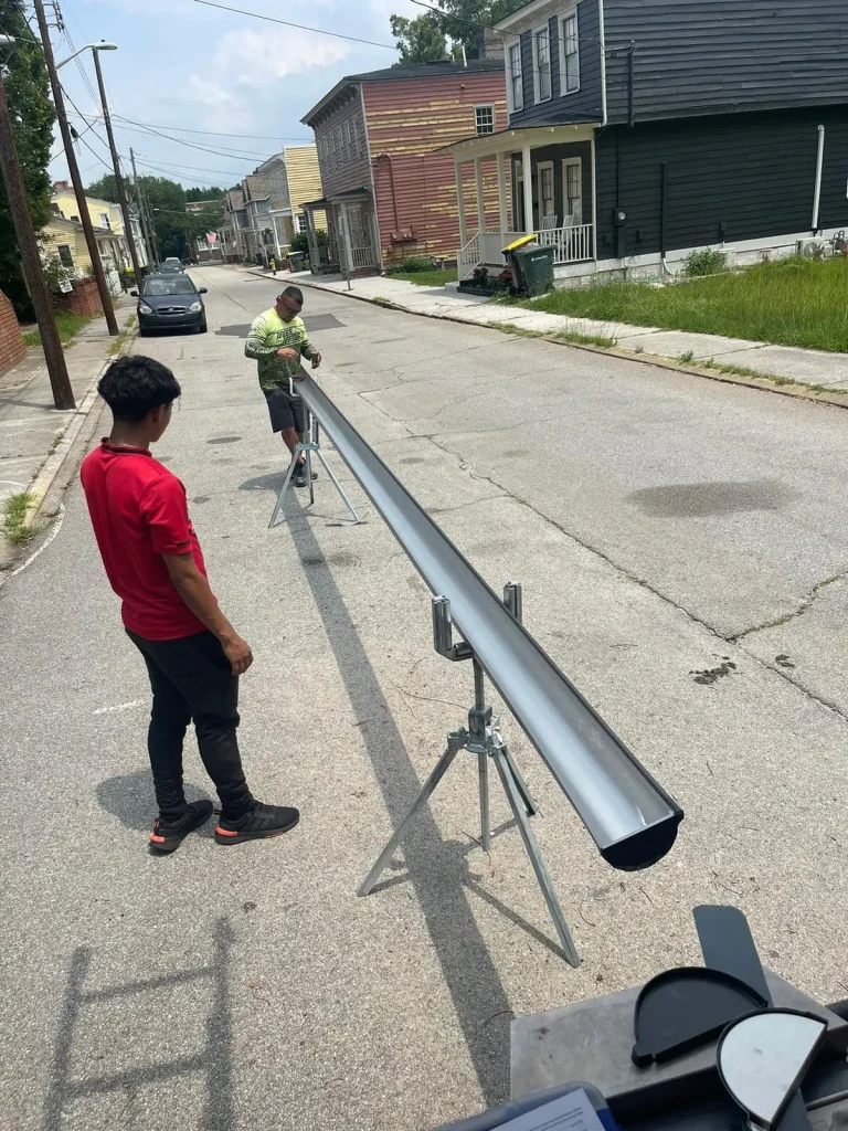 Two gutter installers fitting a metal gutter with supports on a sunny residential street, tools and supplies nearby.