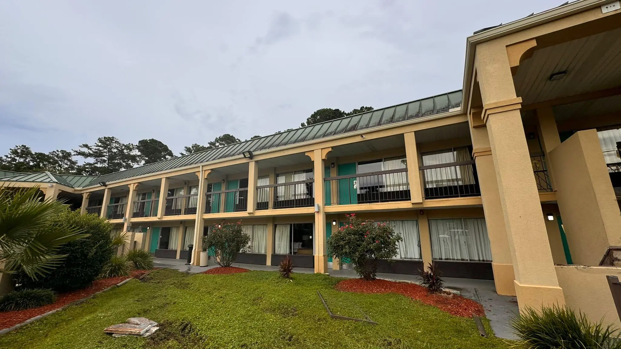 Wide-angle view of a two-story apartment complex featuring a long, sloped green metal roof and tan stucco pillars. A white downspout is visible on a central pillar, draining onto a concrete walkway near a grassy area. The building has a continuous roofline with multiple bays, showing the integration of the gutter system with the second-floor balcony overhangs.