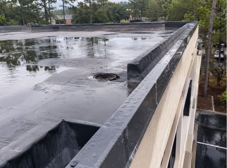 Commercial flat roof in Bluffton, SC after a rainstorm showing standing water near a box gutter drain, with visible flashing details and low-slope roof drainage system under overcast skies.
